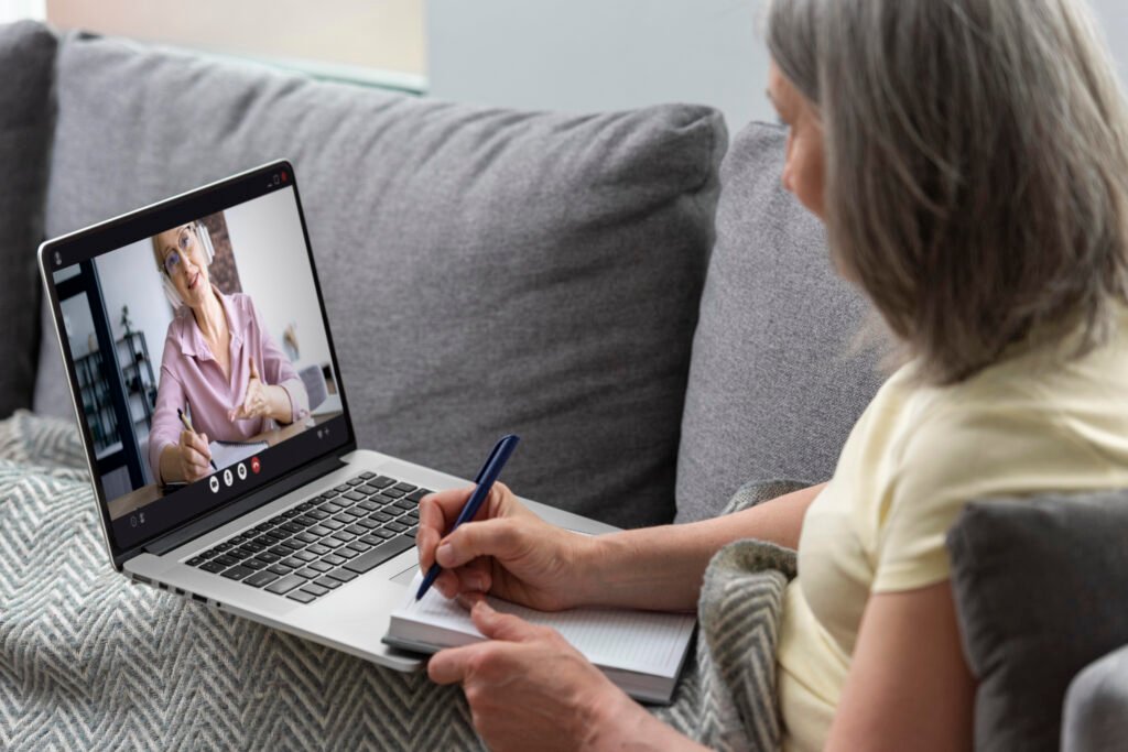 Mulher em uma sala bem iluminada, participando de uma sessão de terapia online com fones de ouvido, olhando para a tela do computador.